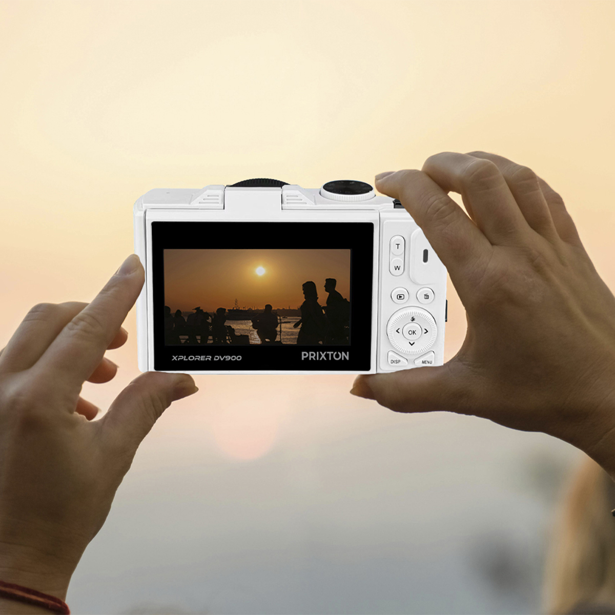 Ręce trzymające biały aparat, wyświetlający zachód słońca na ekranie. Widoczne sylwetki. Tło to pomarańczowe niebo.