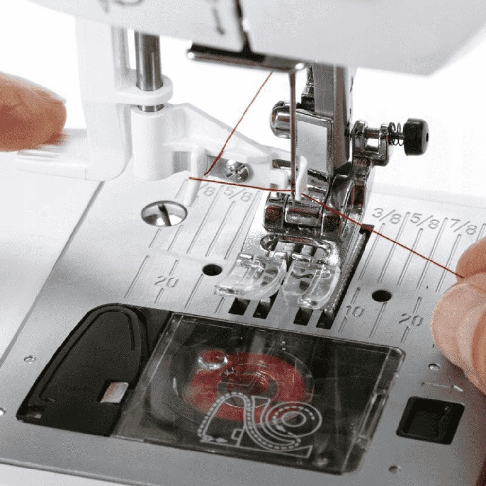 Zbliżenie igły maszyny do szycia z czerwoną nitką. Maszyna jest srebrna i biała. Ręka trzyma nić.