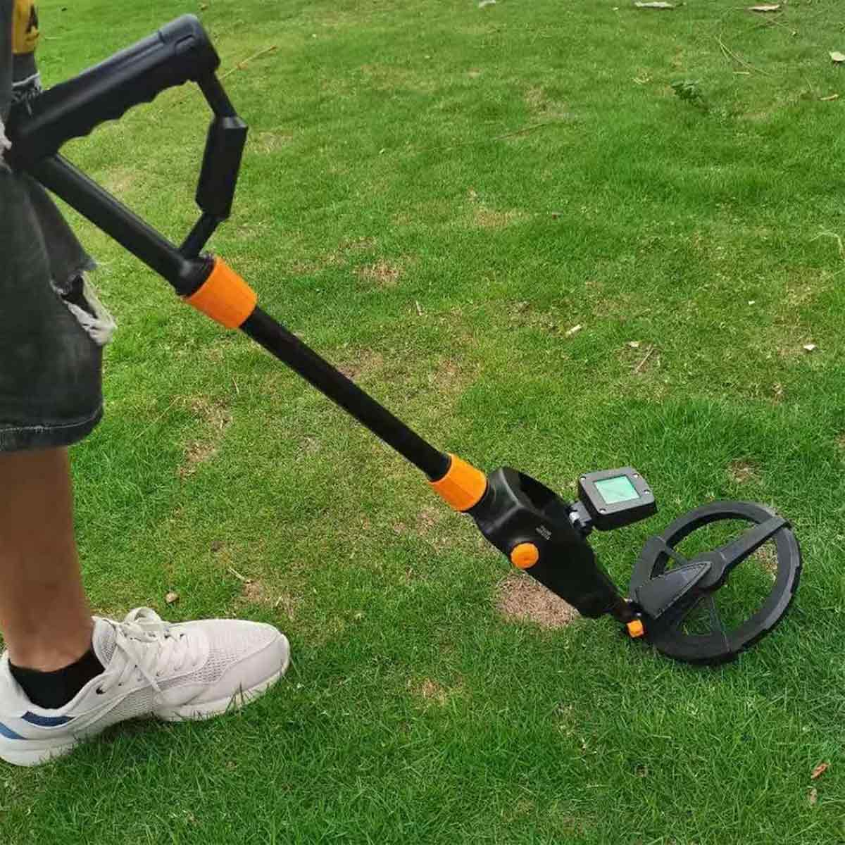 Eine Person benutzt einen Metalldetektor auf grünem Gras. Der Detektor ist schwarz und orange. Die Person trägt weiße Turnschuhe.