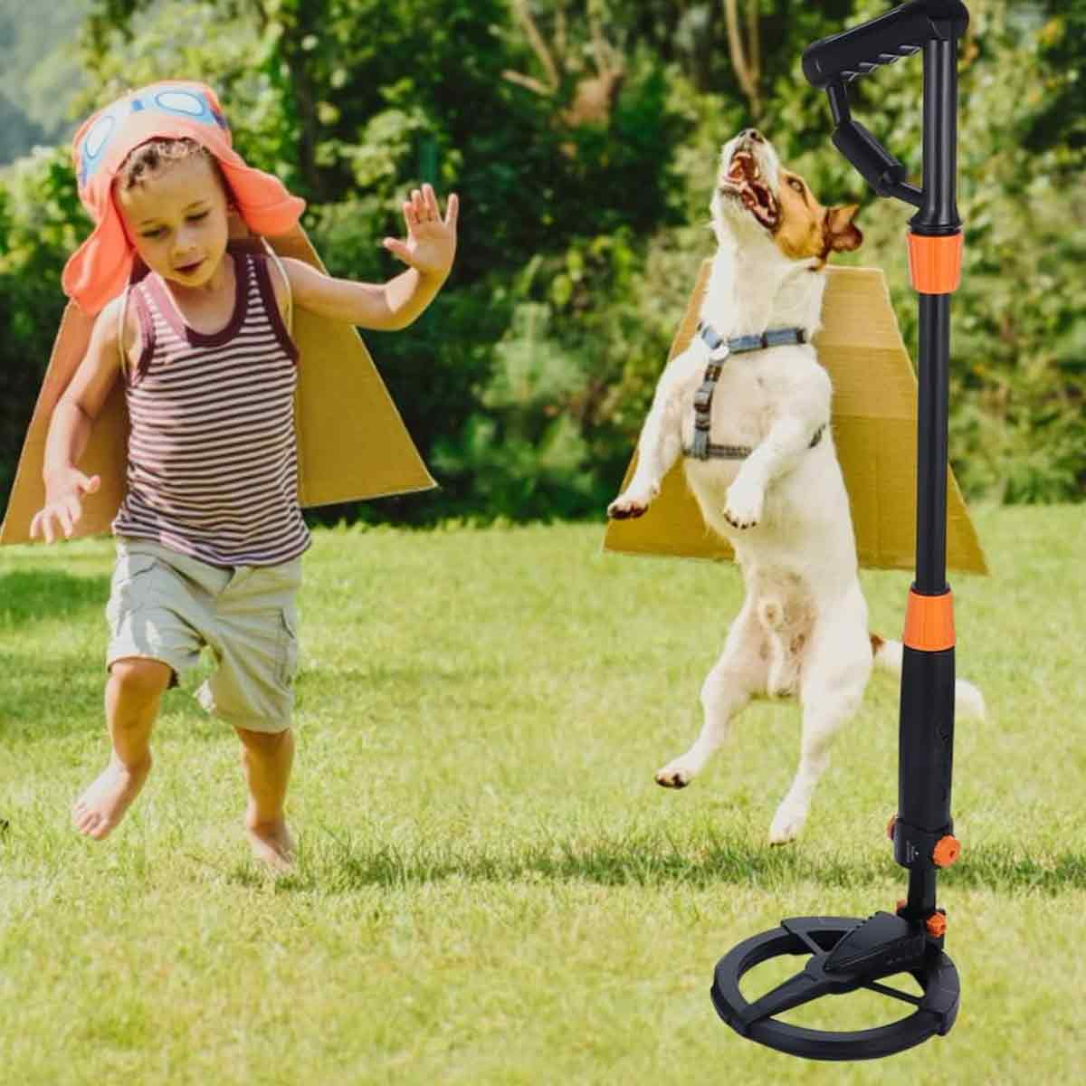 Ein Kind im Kostüm und ein Hund mit Pappflügeln spielen in einem grünen Garten. Ein Metalldetektor steht in der Nähe.