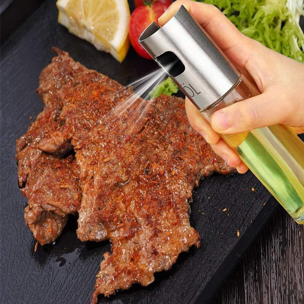 Eine Hand sprüht Öl auf gewürztes Steak. Zitrone, Tomate und Salat sind im Hintergrund. Der Ölsprüher ist aus Metall und Glas.