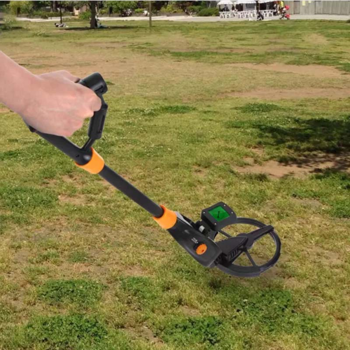 Eine Person benutzt einen Metalldetektor auf einer Wiese. Der Detektor ist schwarz und orange. Im Hintergrund sind Bäume und Menschen.