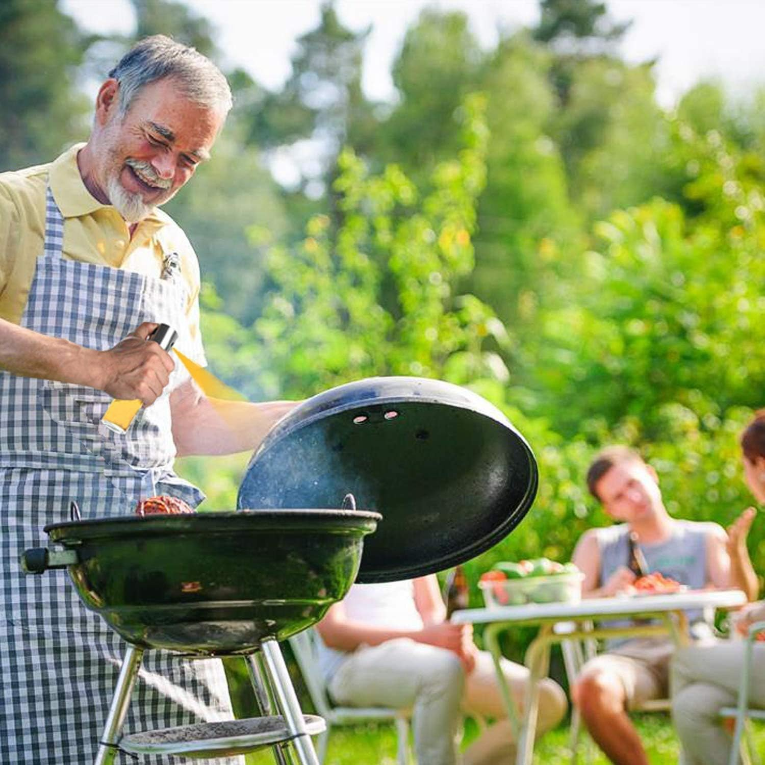 Ein Mann lächelt beim Grillen. Er trägt eine Schürze und hält ein Spray. Im Hintergrund sitzen Leute an einem Tisch.