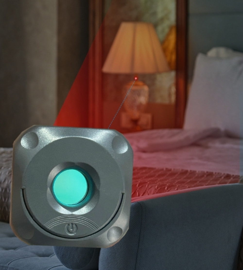 Ein silbernes Gerät mit grünem Zentrum ist auf ein Bett gerichtet. Ein roter Laserstrahl erstreckt sich zu einer Lampe.