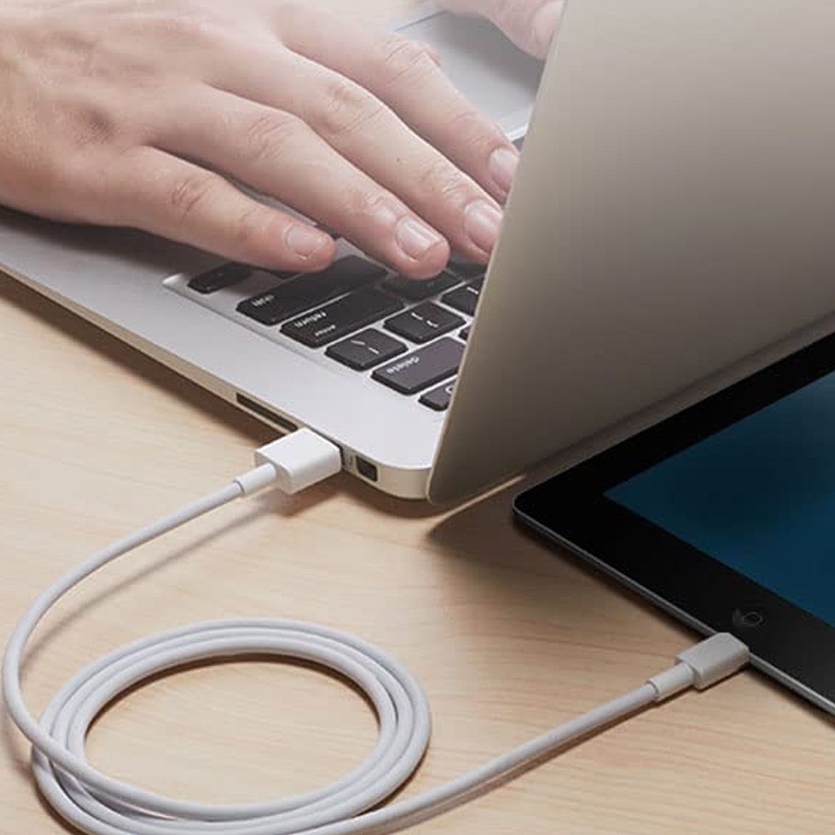 Eine Hand tippt auf einem Laptop. Ein weißes Kabel verbindet Laptop und Tablet auf Holz.