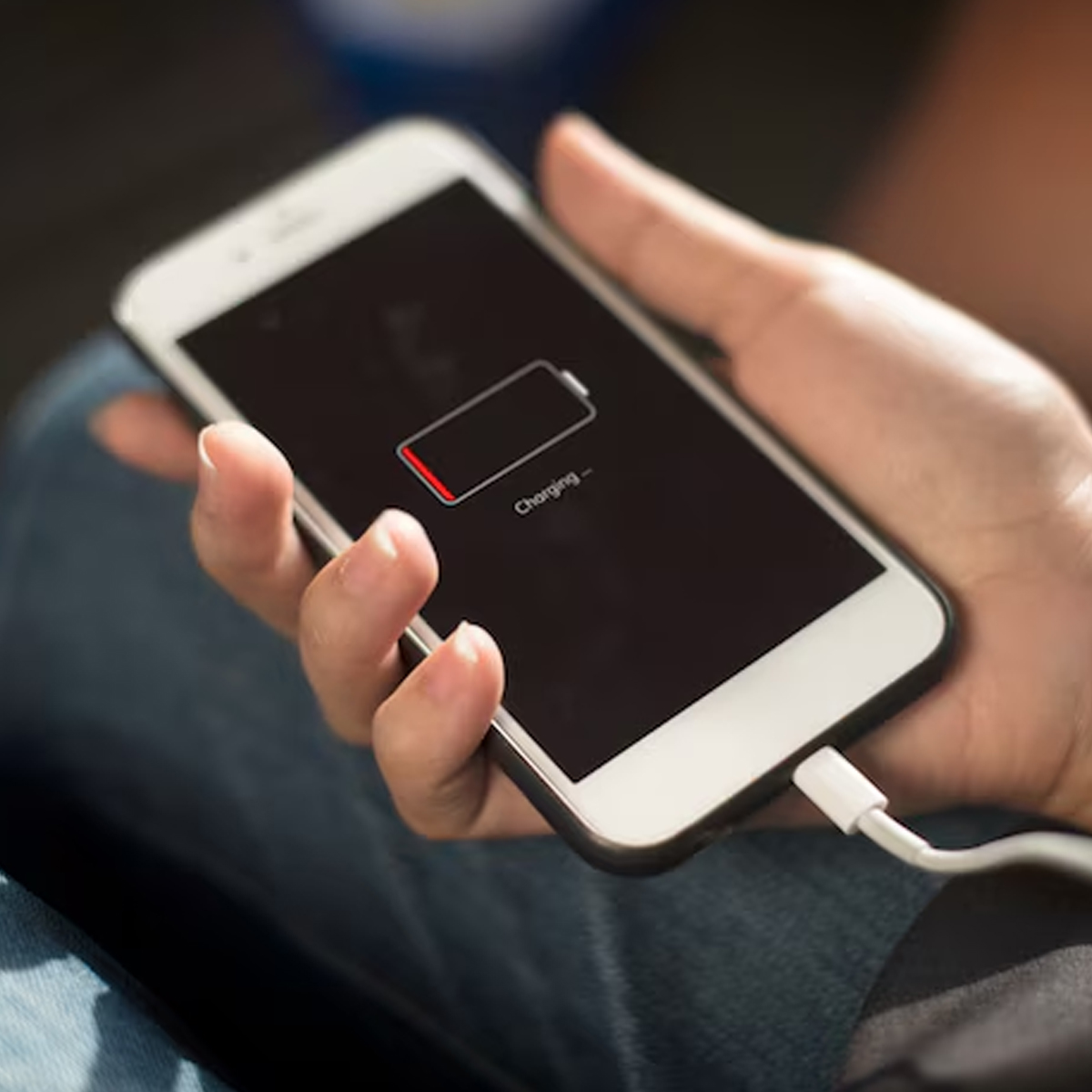 Eine Hand hält ein weißes Smartphone mit eingestecktem Ladekabel. Der Bildschirm zeigt ein Symbol für schwache Batterie und das Wort 'Charging'.
