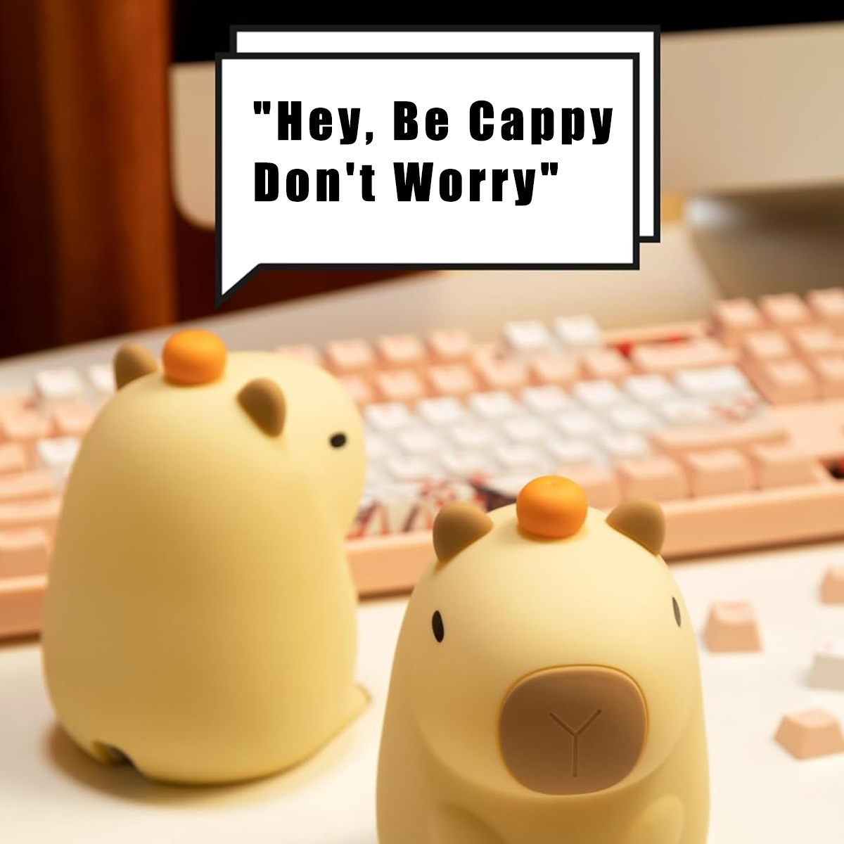 Zwei gelbe Capybara-Figuren, eine davon im Fokus. Eine Sprechblase sagt: 'Hey, Be Cappy Don't Worry'. Im Hintergrund eine rosa Tastatur.