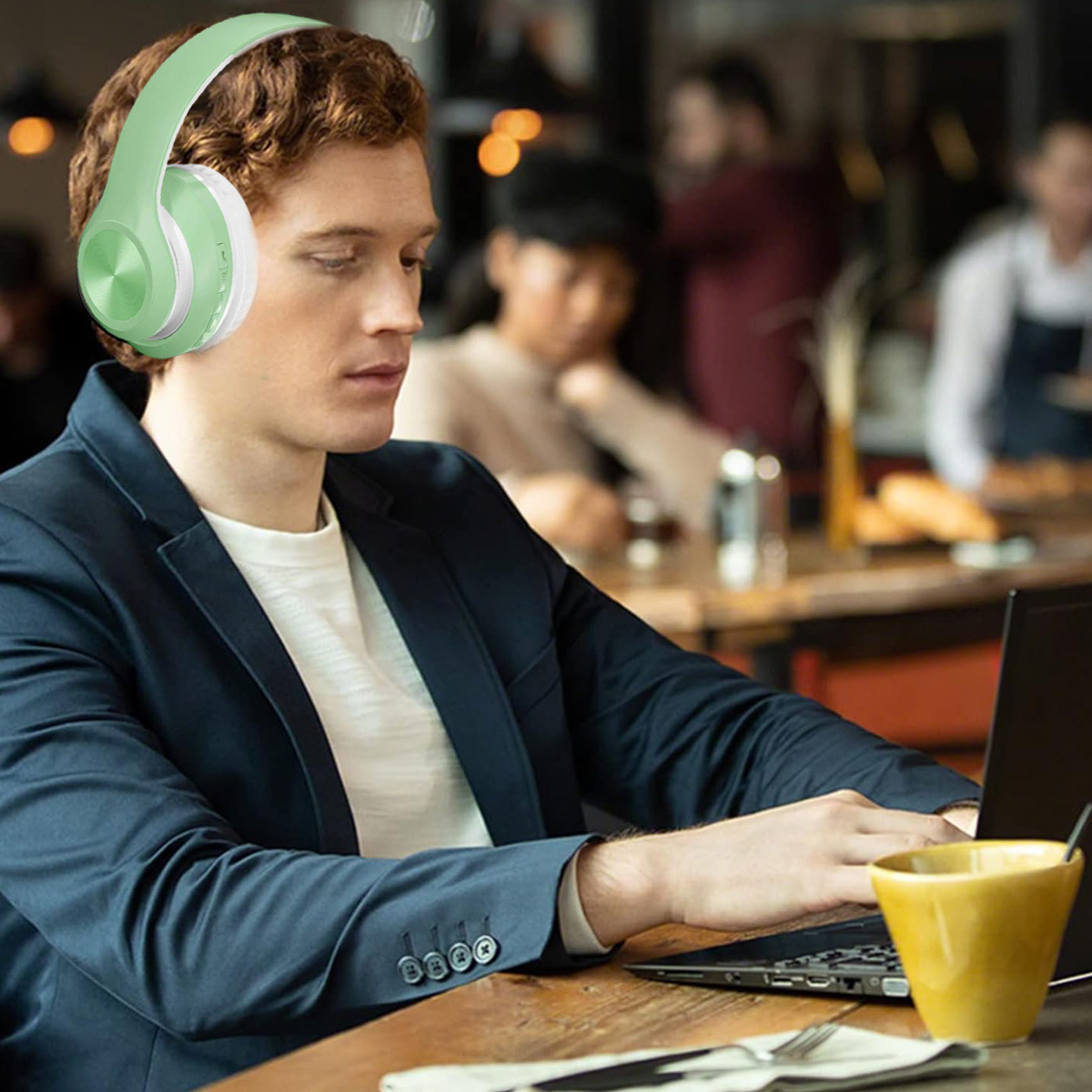 Ein Mann mit roten Haaren und Kopfhörern benutzt in einem Café einen Laptop. Auf dem Tisch stehen eine gelbe Tasse und Besteck.