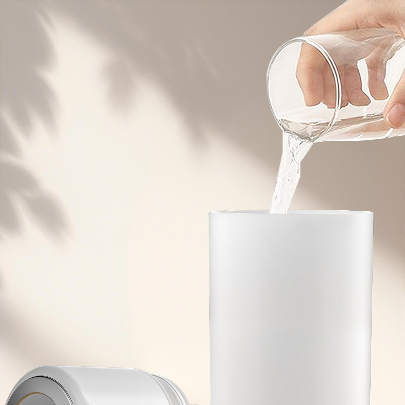 Eine Hand gießt Wasser aus einem Glas in einen weißen Zylinder. Der Hintergrund ist hellbeige mit Schatten.