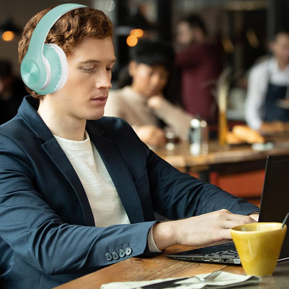 Ein Mann mit Kopfhörern benutzt in einem Café einen Laptop. Auf dem Tisch stehen eine gelbe Tasse und Besteck. Andere Personen sind im Hintergrund unscharf.