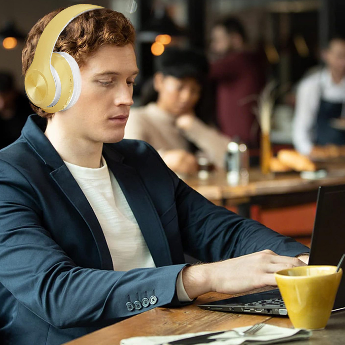Ein Mann mit Kopfhörern arbeitet in einem Café an einem Laptop. Er trägt einen marineblauen Blazer und auf dem Tisch steht eine gelbe Tasse.