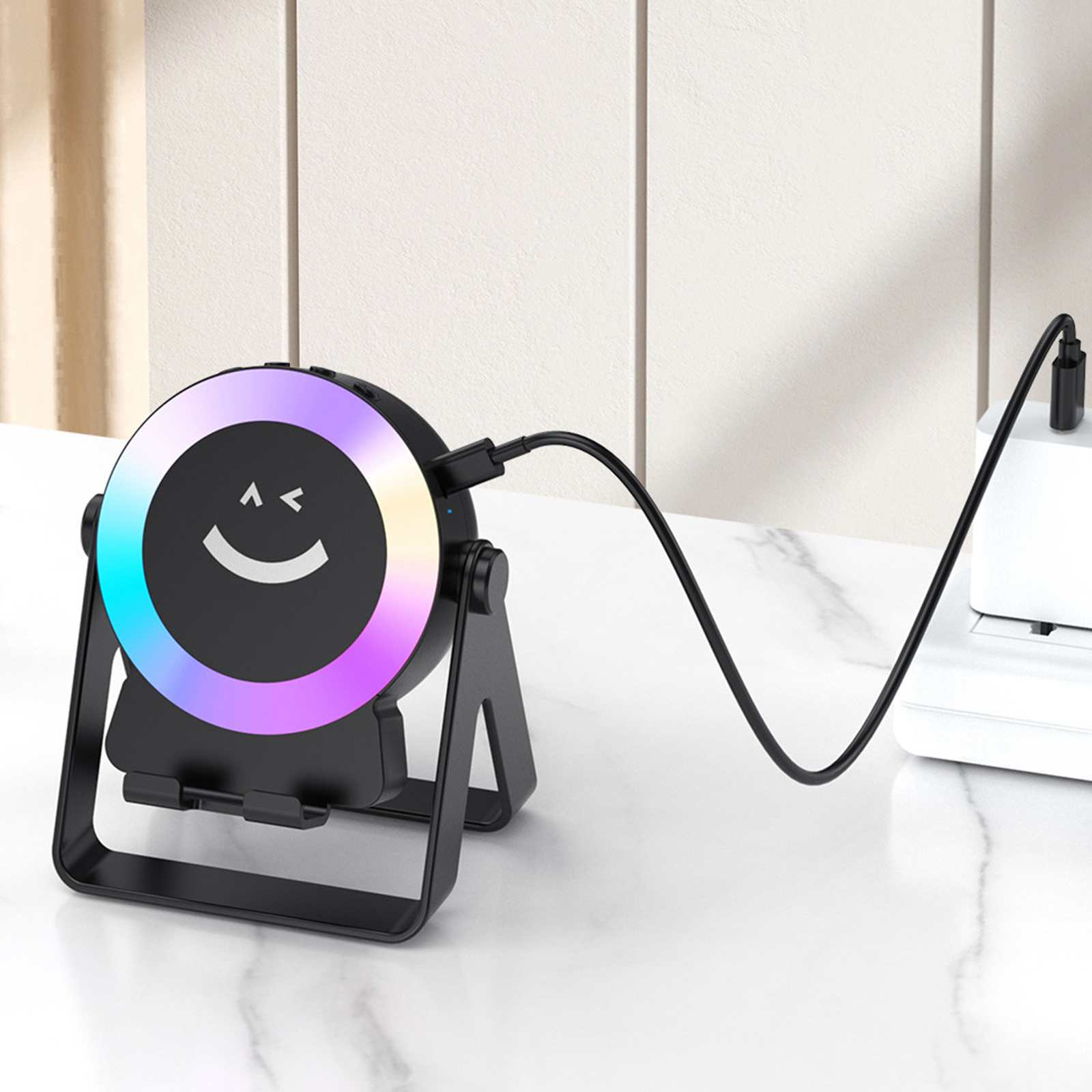 Ein schwarzes Gerät mit Regenbogenlichtring, Smiley-Gesicht und Kabel, das auf einem Ständer ruht. An eine weiße Box angeschlossen.