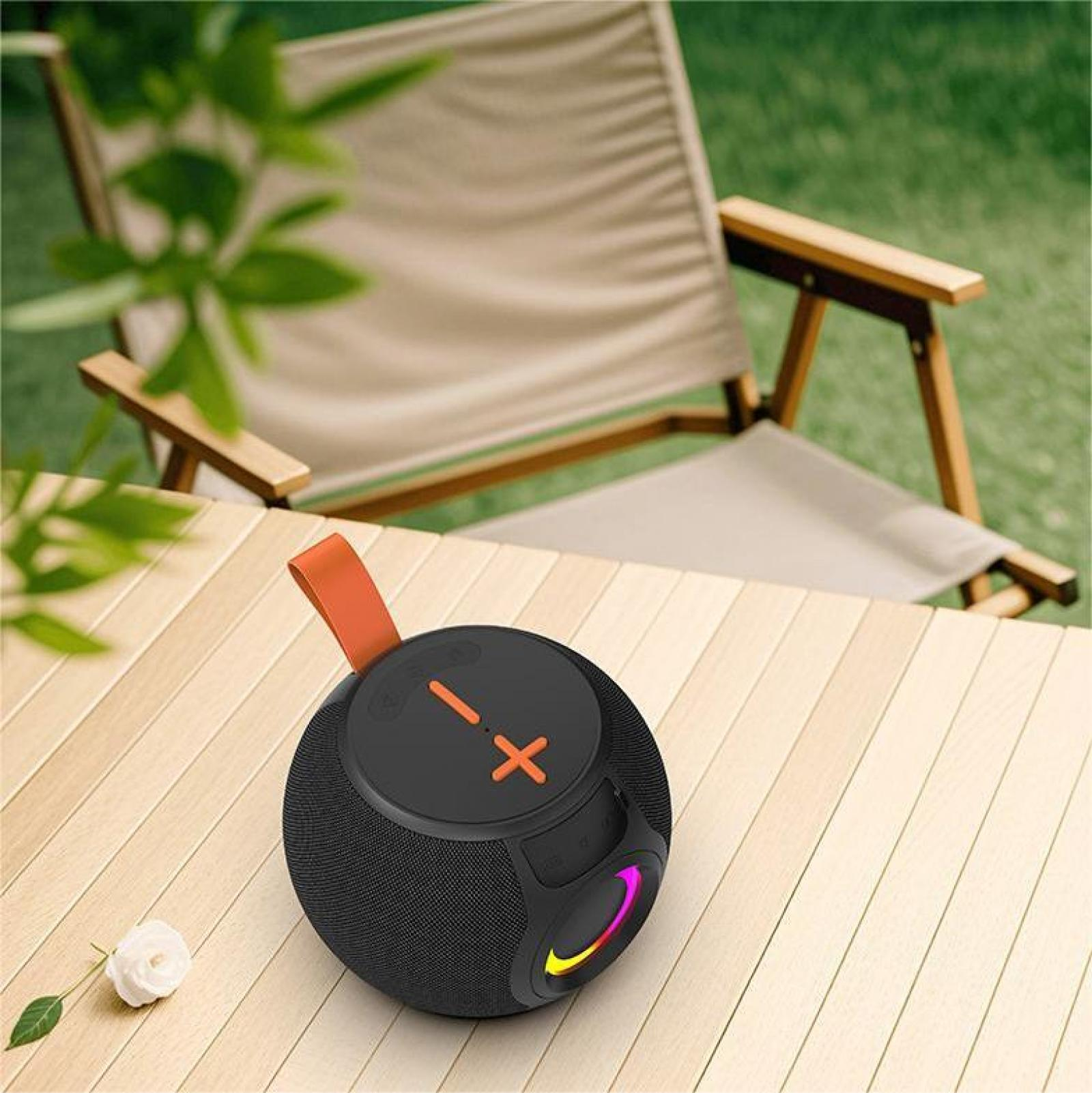 Ein schwarzer Lautsprecher mit orangefarbenem Griff steht auf einem Holztisch. Eine weiße Blume und ein Stuhl sind im Hintergrund.