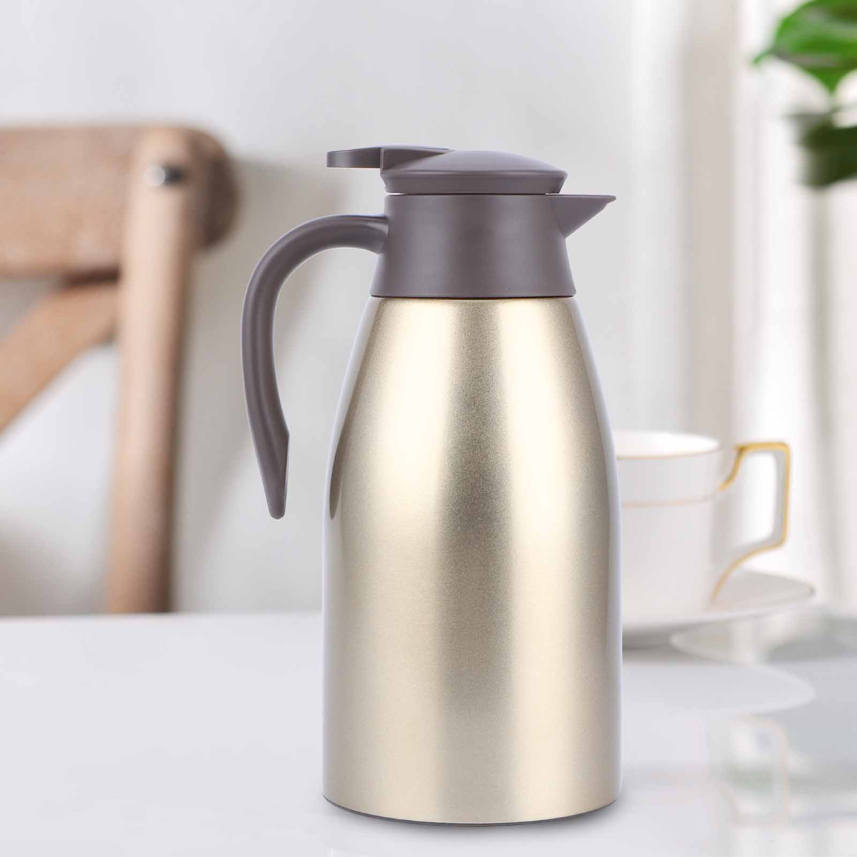 Eine goldene Thermoskanne mit braunem Griff und Deckel steht auf einem weißen Tisch. Im Hintergrund sind eine Tasse und ein Stuhl zu sehen.
