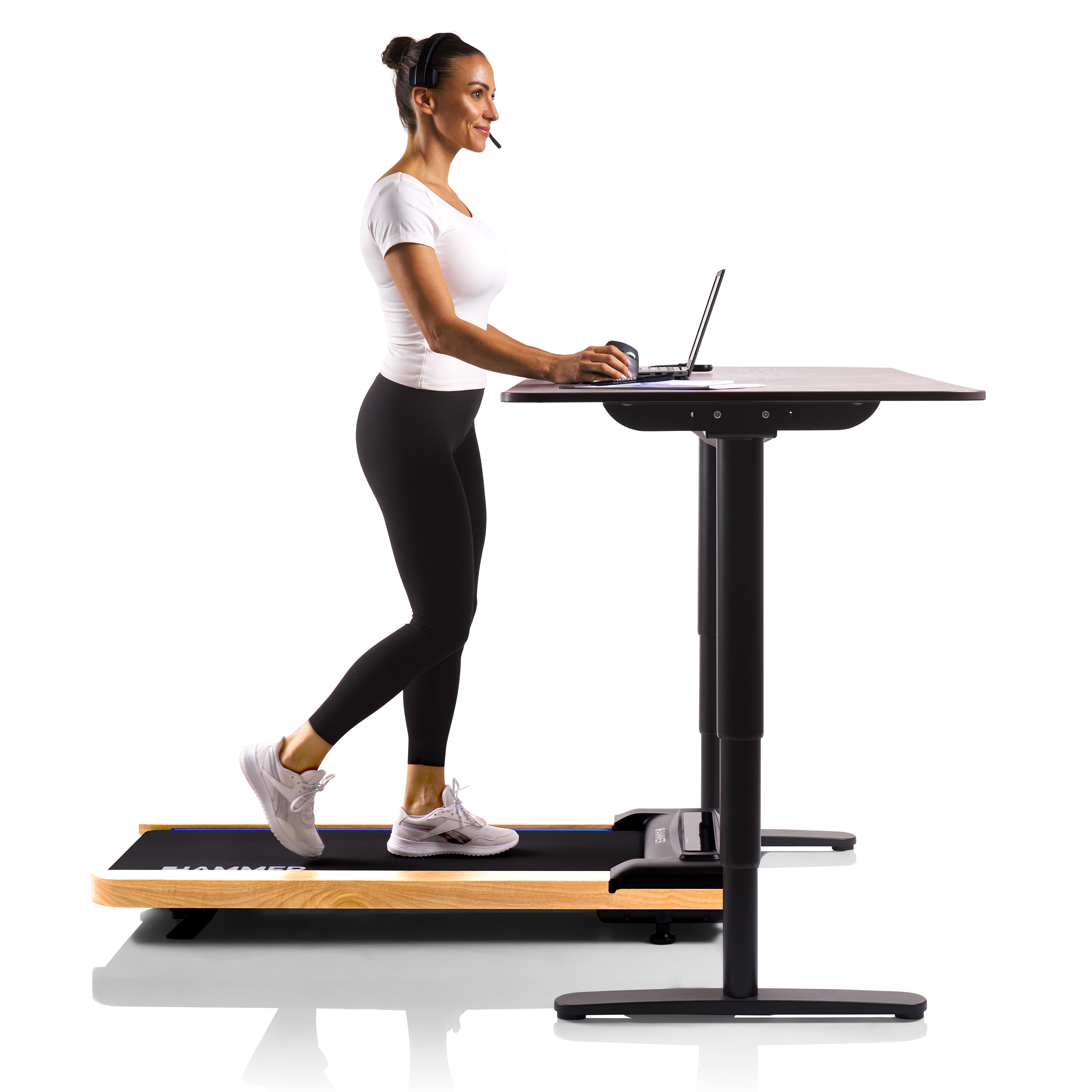 Frau, die auf einem Laufband-Schreibtisch geht, mit Headset, arbeitet an einem Laptop. Schwarze Leggings und weißes Hemd.