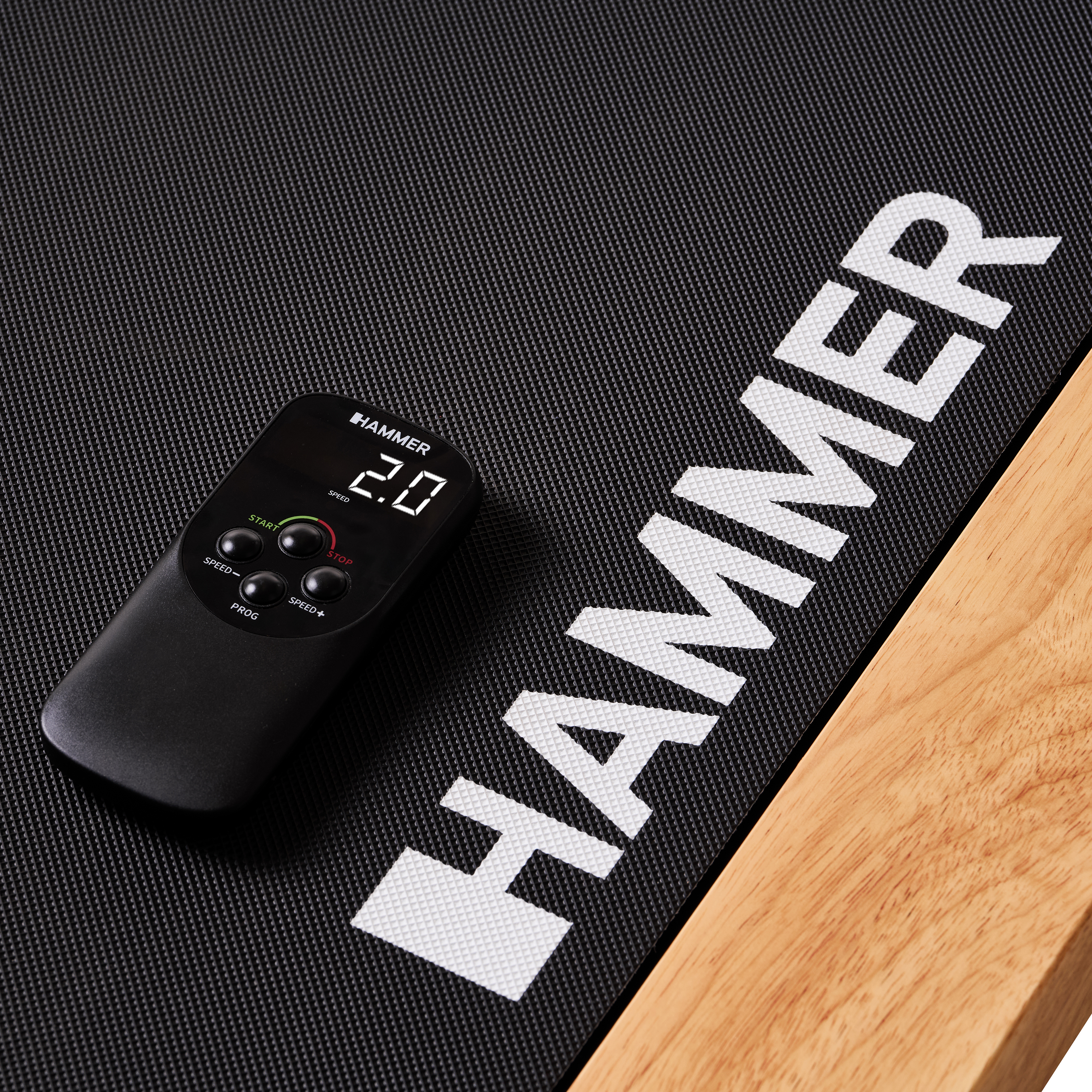 Schwarze Laufbandoberfläche mit Fernbedienung. Hammer-Logo und Geschwindigkeitsanzeige. Holzseite rechts.