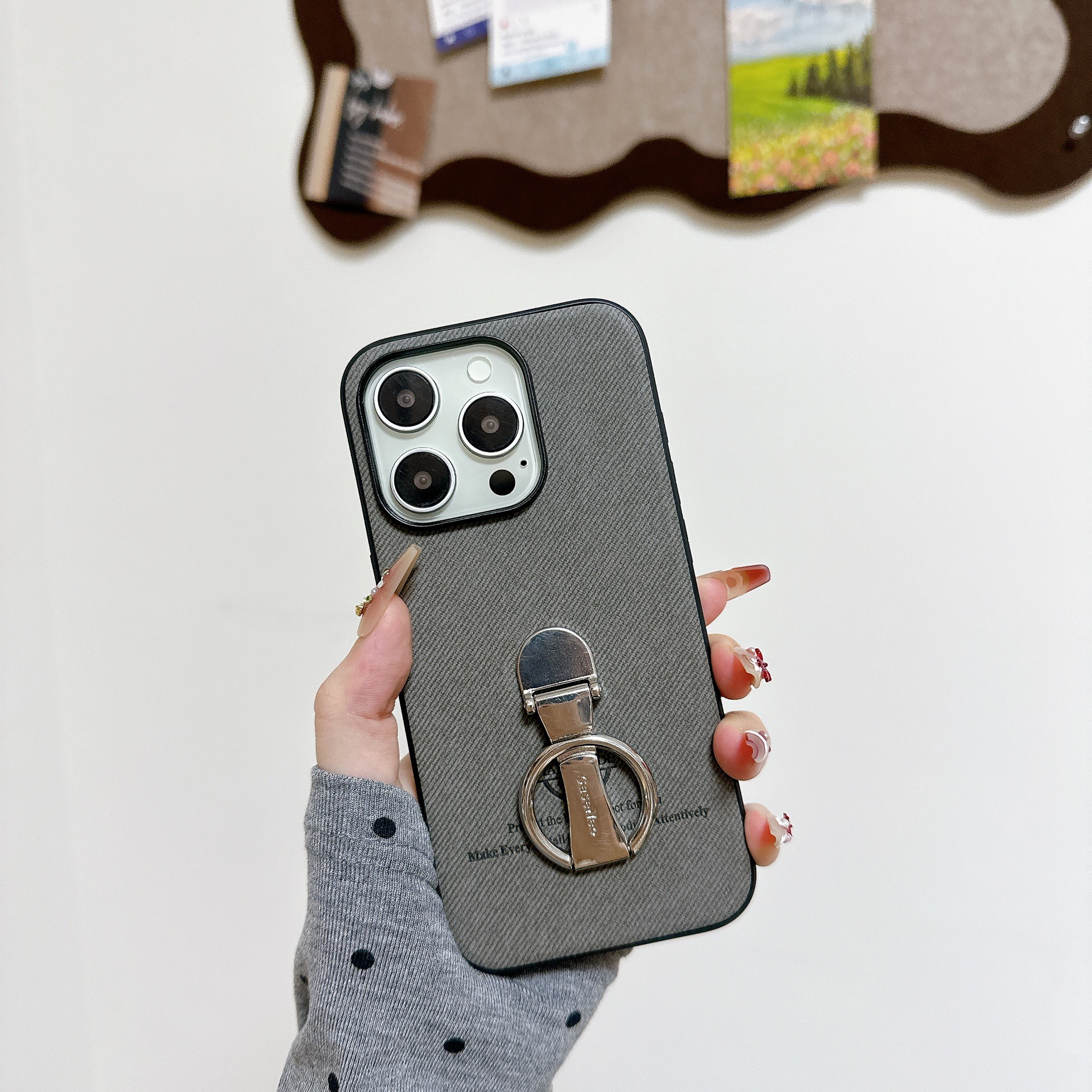 Una mano tiene una custodia grigia per telefono con un anello argentato. Sullo sfondo una bacheca con piccole immagini.