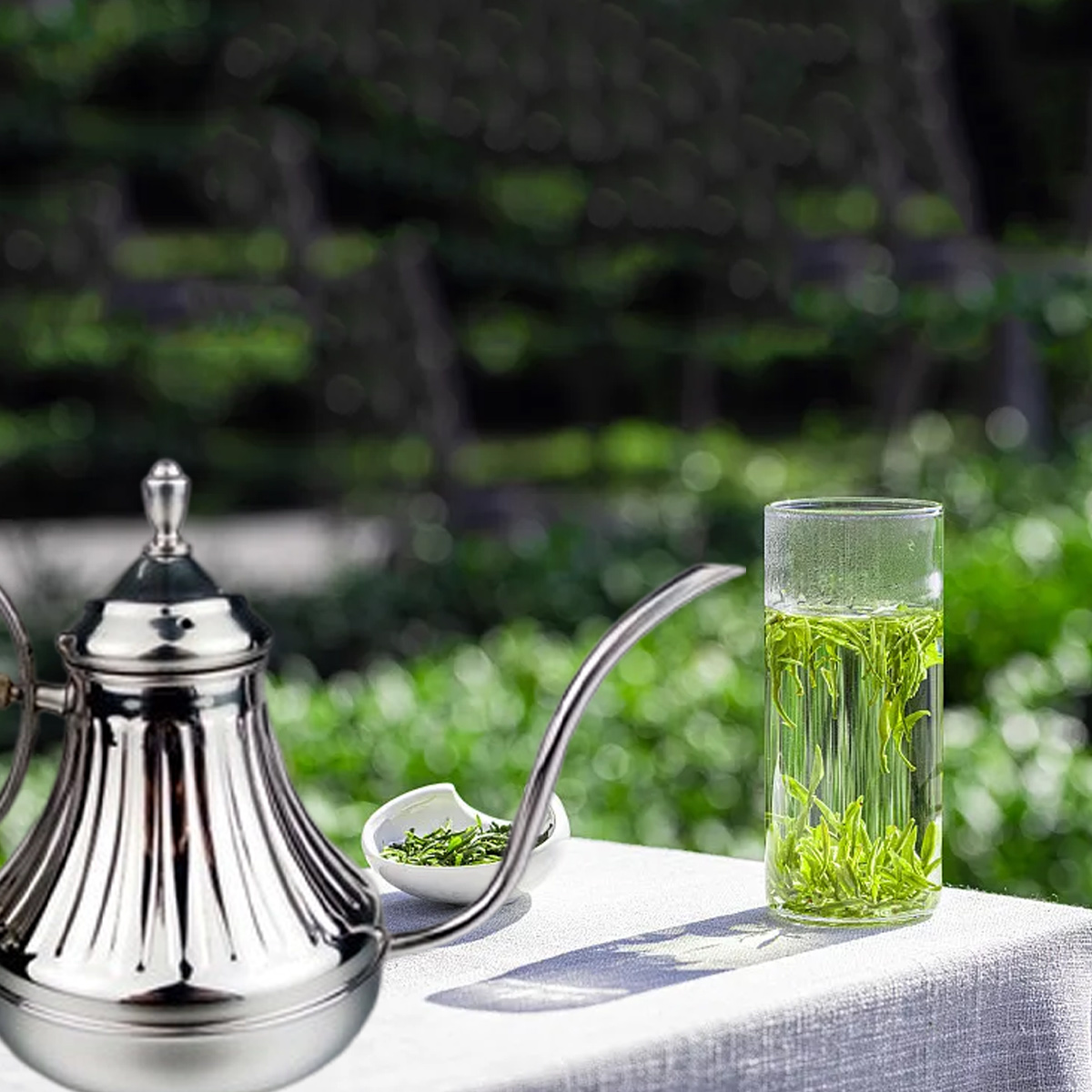 Eine silberne Teekanne, ein Glas Tee und eine kleine Schüssel mit Teeblättern stehen auf einem weißen Tuch. Grünes Laub im Hintergrund.