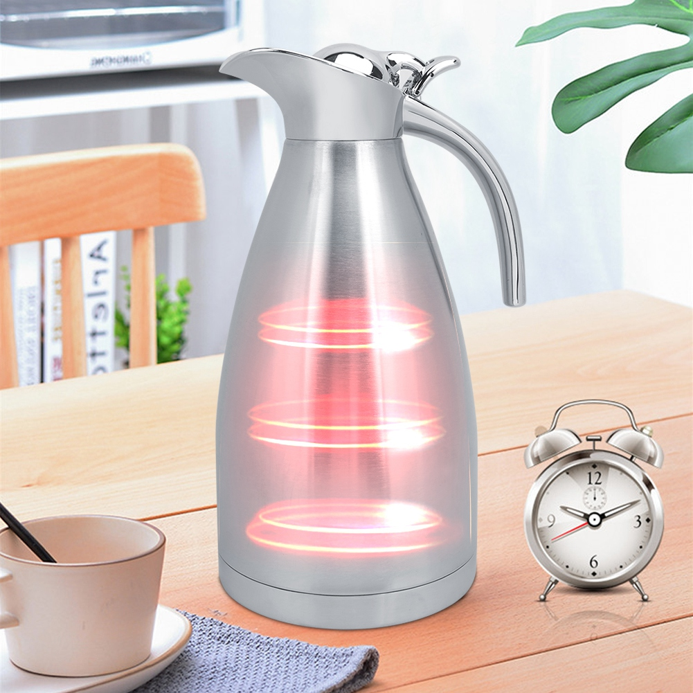 Eine silberne Isolierkanne mit roten Glühelementen steht auf einem Holztisch neben einer Uhr und einer Tasse.