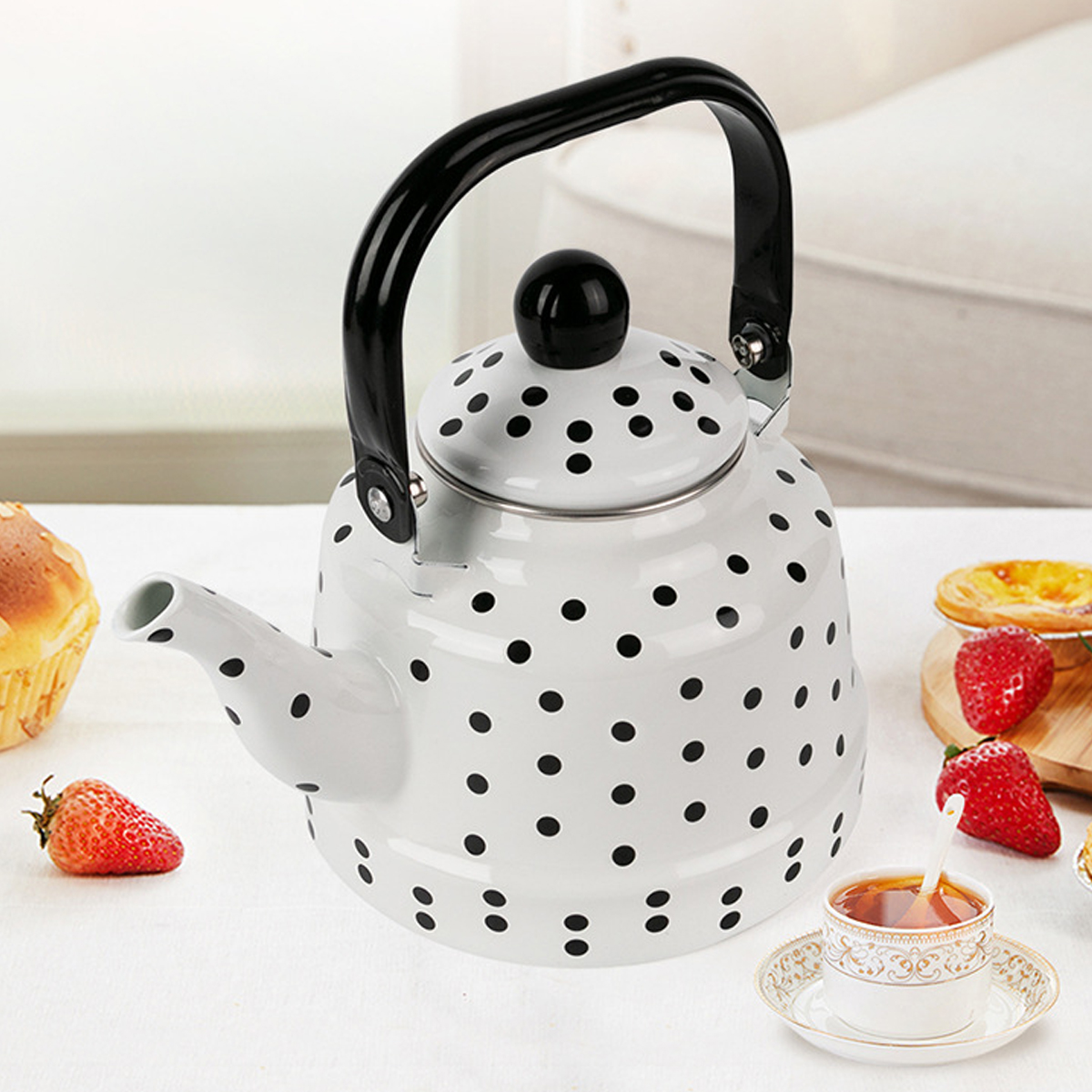 Eine weiße Teekanne mit schwarzen Punkten und einem schwarzen Griff steht auf einem weißen Tisch. Erdbeeren und Gebäck umgeben eine Teetasse.