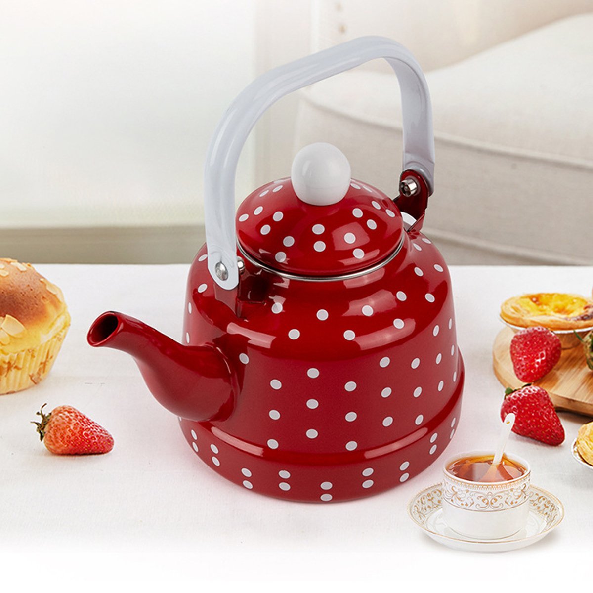 Ein roter Polka-Dot-Kessel mit weißem Griff steht auf einem weißen Tisch. Erdbeeren, Kuchen und Teetasse sind ebenfalls zu sehen.