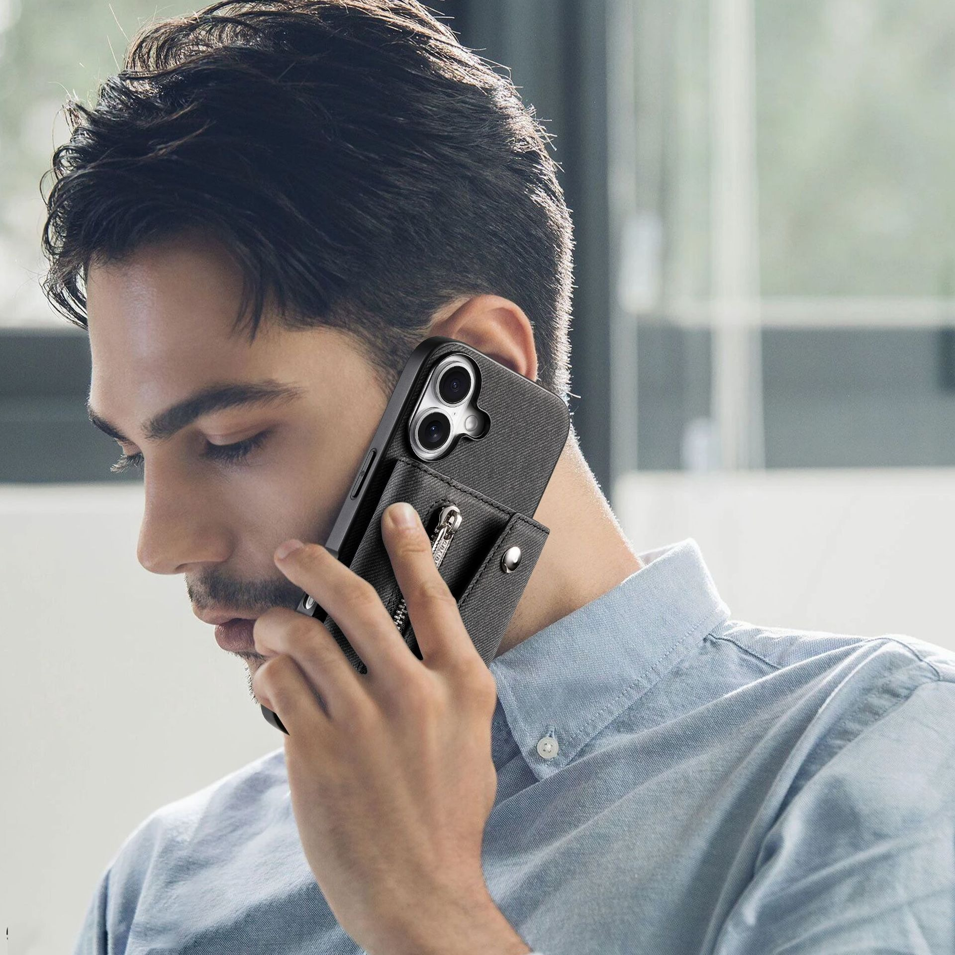 Un uomo con una camicia blu tiene in mano un telefono nero con custodia. Guarda in basso mentre parla al telefono.
