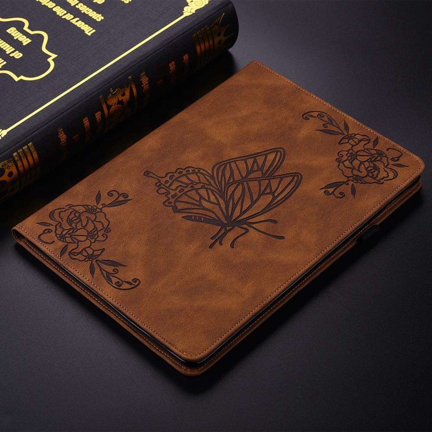 Braune Leder-Buchhülle mit Schmetterlings- und Blumendesign. Ein dunkles Buch und eine schwarze Oberfläche sind ebenfalls sichtbar.