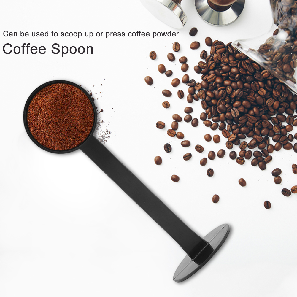 Ein schwarzer Kaffeelöffel mit braunem Kaffeepulver, umgeben von Kaffeebohnen auf weißer Oberfläche.
