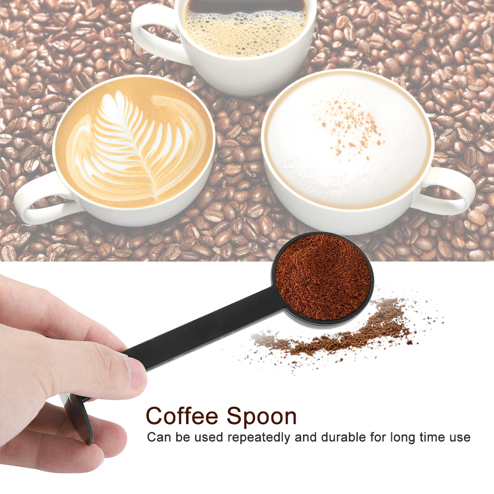 Eine Hand hält einen schwarzen Kaffeelöffel mit Kaffeepulver. Im Hintergrund sind drei Kaffeetassen und Bohnen zu sehen.