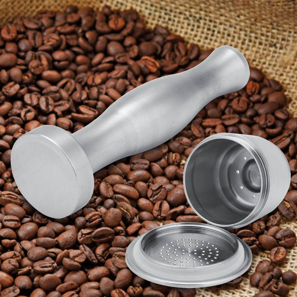 Ein Kaffeestempel und zwei zerlegte Kaffeekapseln liegen auf einem Bett aus Kaffeebohnen. Der Hintergrund ist ein Jutesack.