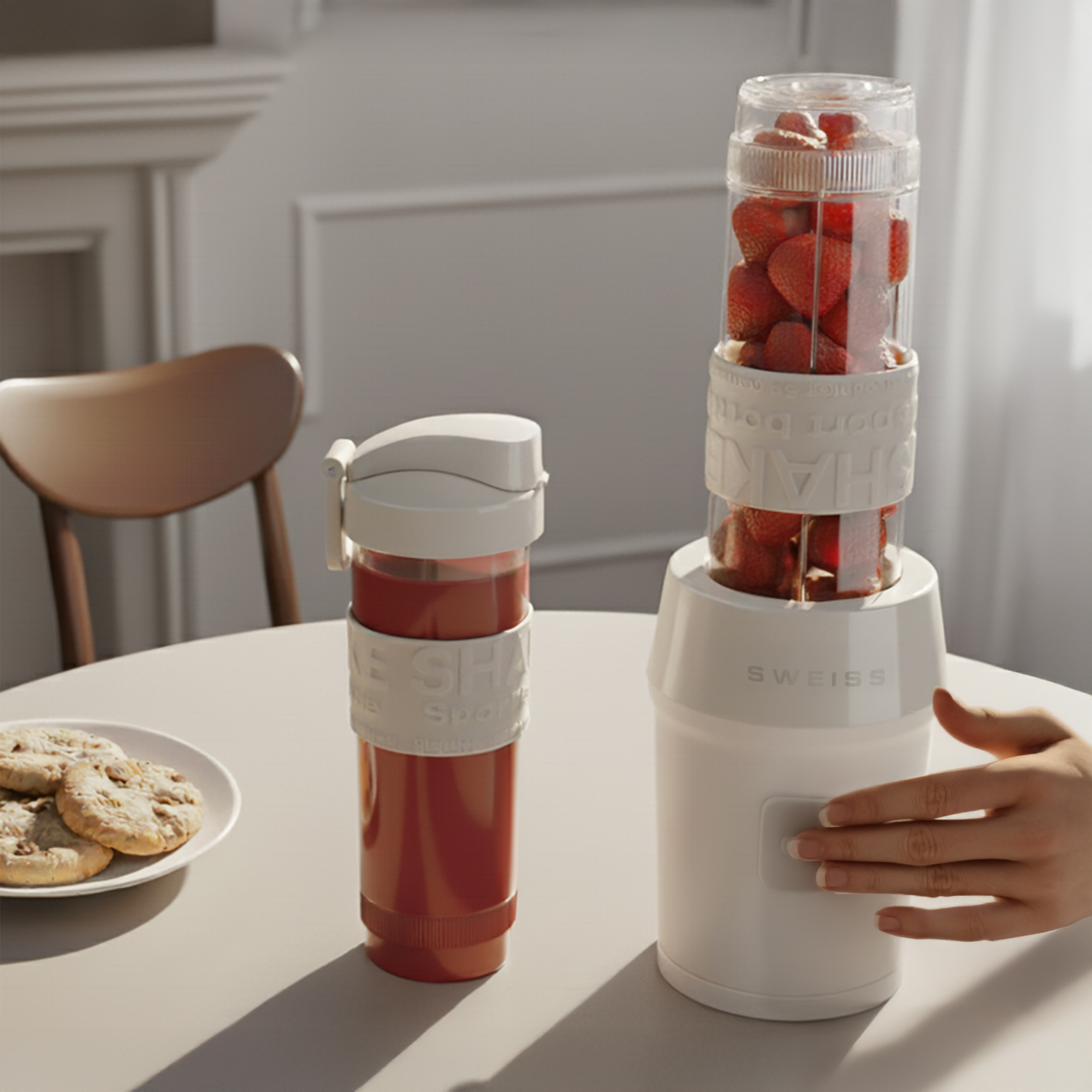 Una licuadora con fresas, una coctelera vacía y galletas en una mesa. Una mano está a punto de presionar el botón de la licuadora.