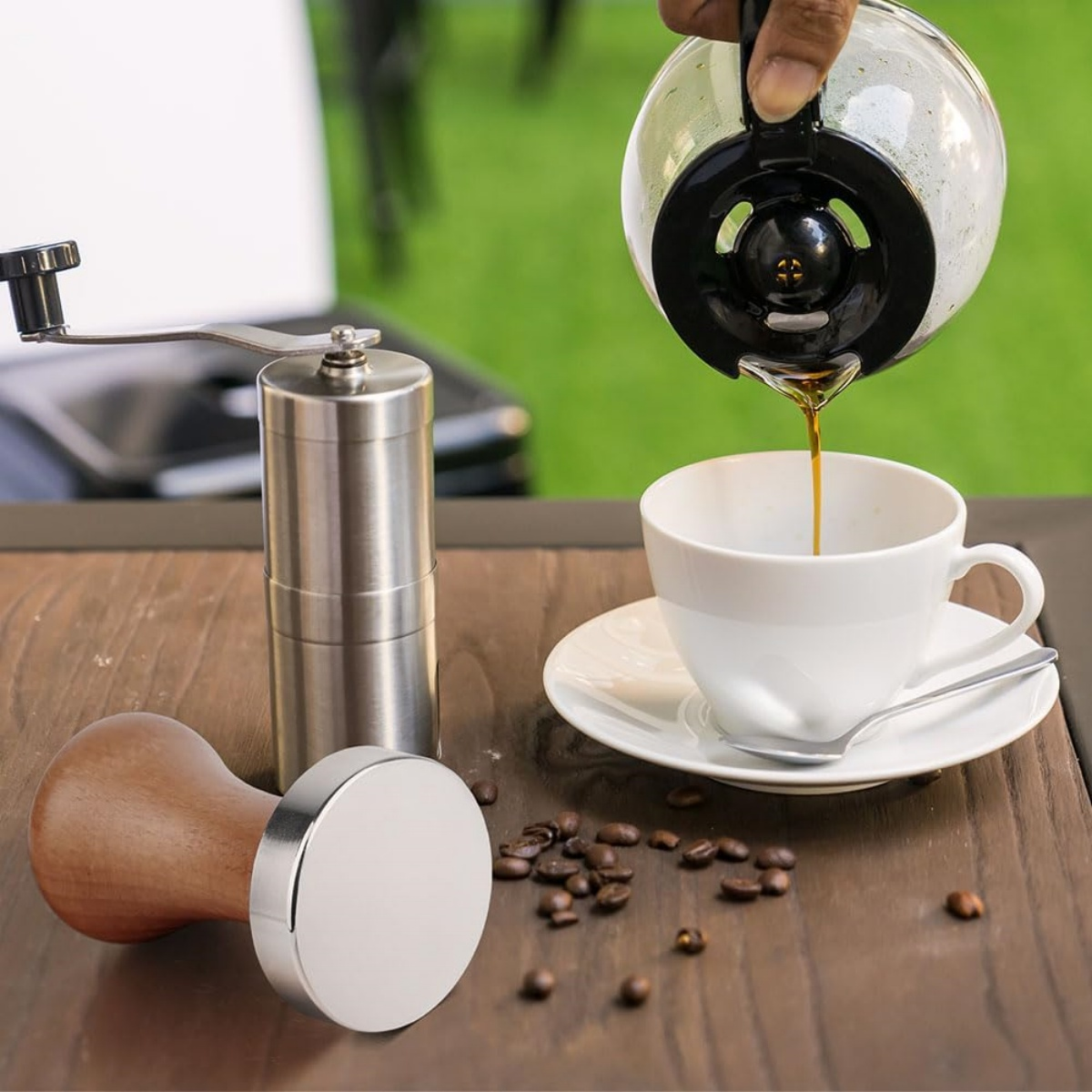 Eine Person gießt Kaffee in eine weiße Tasse, mit Kaffeebohnen, einer Mühle und einem Tamper auf einem Holztisch.