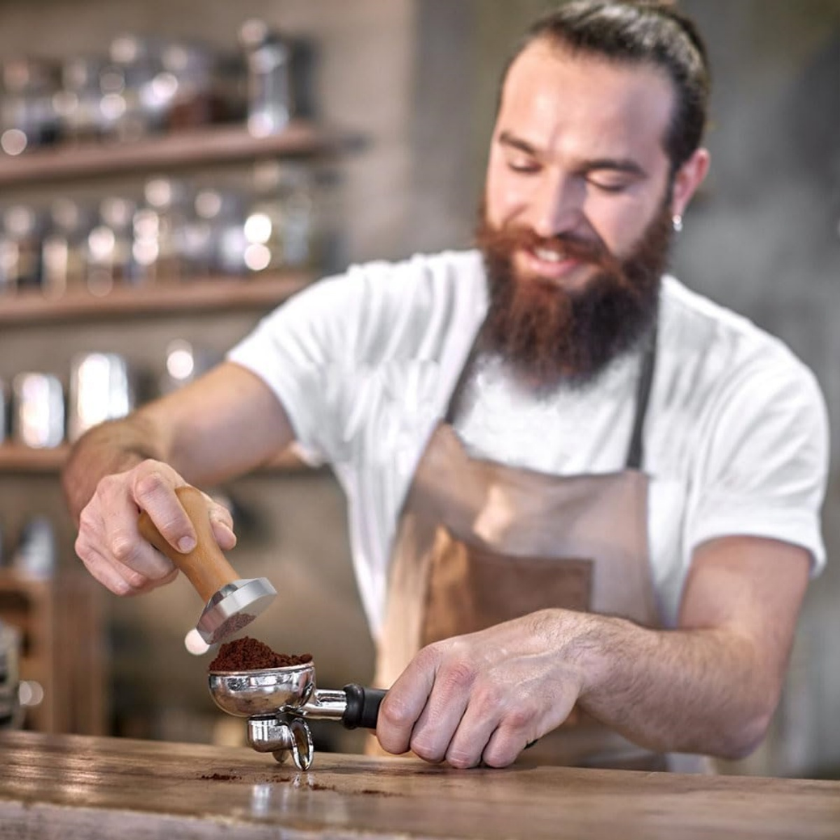 Ein Barista drückt Kaffeepulver mit einem Tamper in einen Siebträger. Er trägt ein weißes Hemd und eine Schürze. Im Hintergrund sind Regale.
