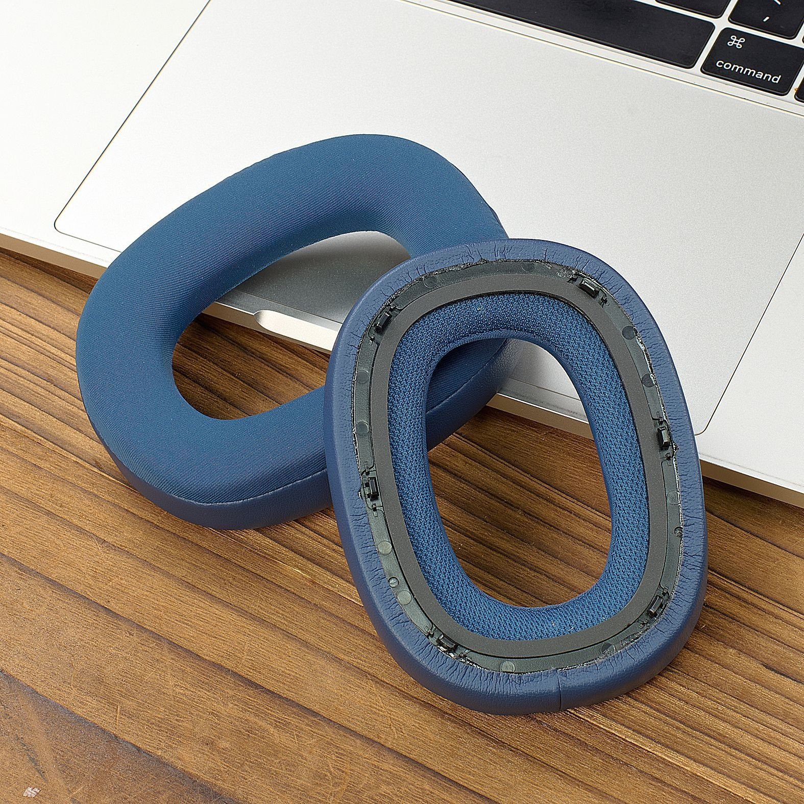 Two blue headphone ear cups on a wooden surface, near a laptop keyboard. One cup is open, the other is closed.