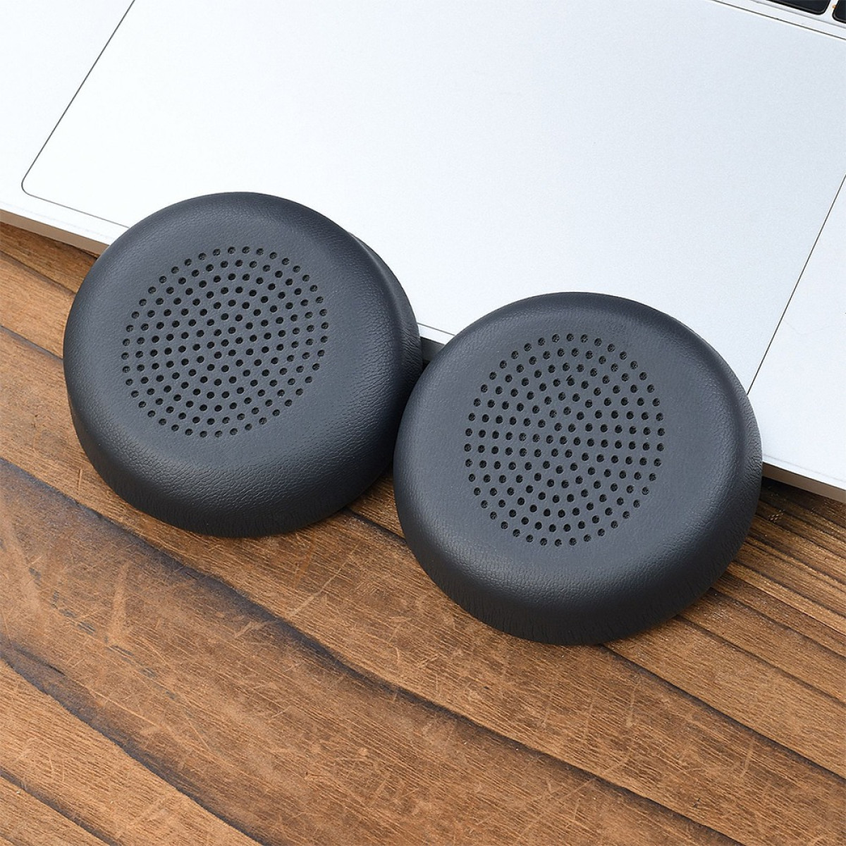 Due cuscinetti auricolari per cuffie grigio scuro su una superficie di legno, parzialmente su un laptop.