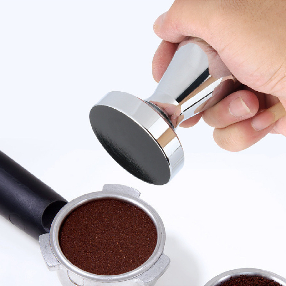 Eine Hand hält einen Chrom-Kaffeestampfer über einem mit Kaffeepulver gefüllten Siebträger auf weißem Hintergrund.