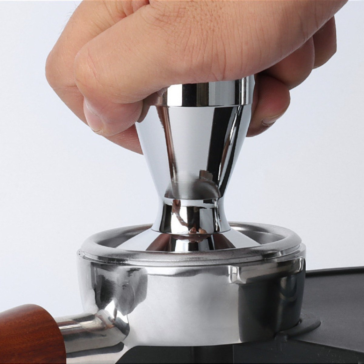 Eine Hand drückt auf einen glänzenden Metallstampfer, der zum Verdichten von Kaffeepulver verwendet wird. Der Tamper befindet sich über einem Siebträger.