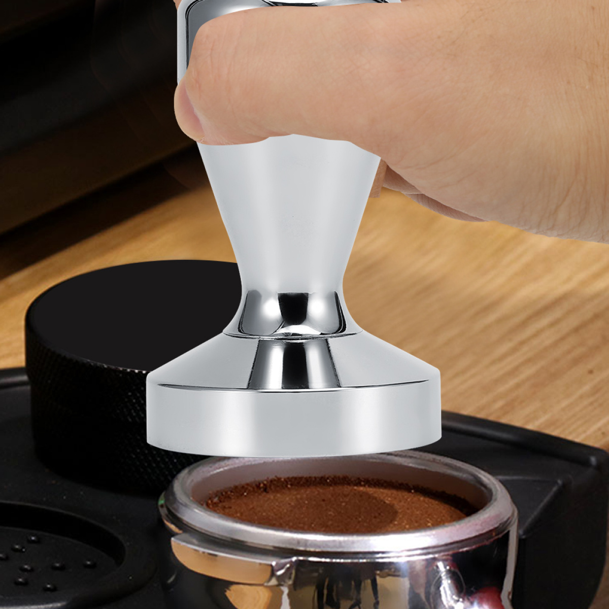 Eine Hand drückt einen silbernen Kaffeestempel auf gemahlenen Kaffee in einem Siebträger. Ein schwarzes Objekt und Holz sind auch zu sehen.