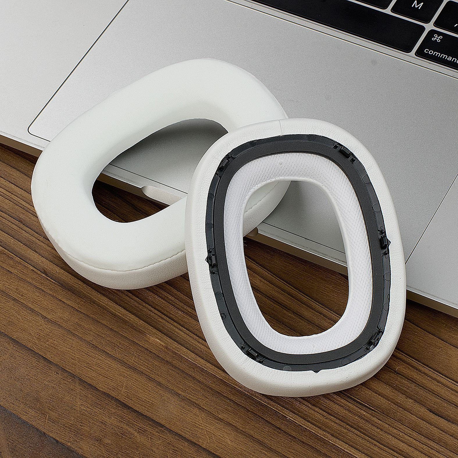 Due cuscinetti auricolari bianchi per cuffie giacciono su una superficie di legno e un laptop. Un cuscinetto è smontato.