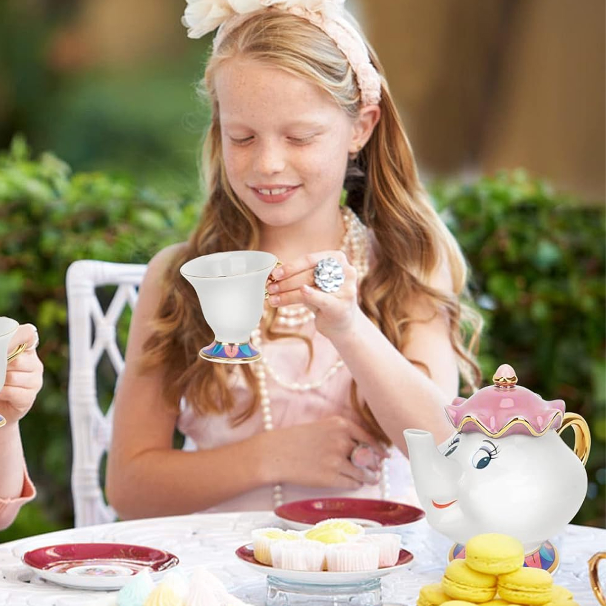Ein Mädchen hält eine Teetasse bei einer Teeparty. Auf dem Tisch stehen eine Teekanne, Makronen und Cupcakes. Ein weißer Stuhl und grüne Büsche sind zu sehen.