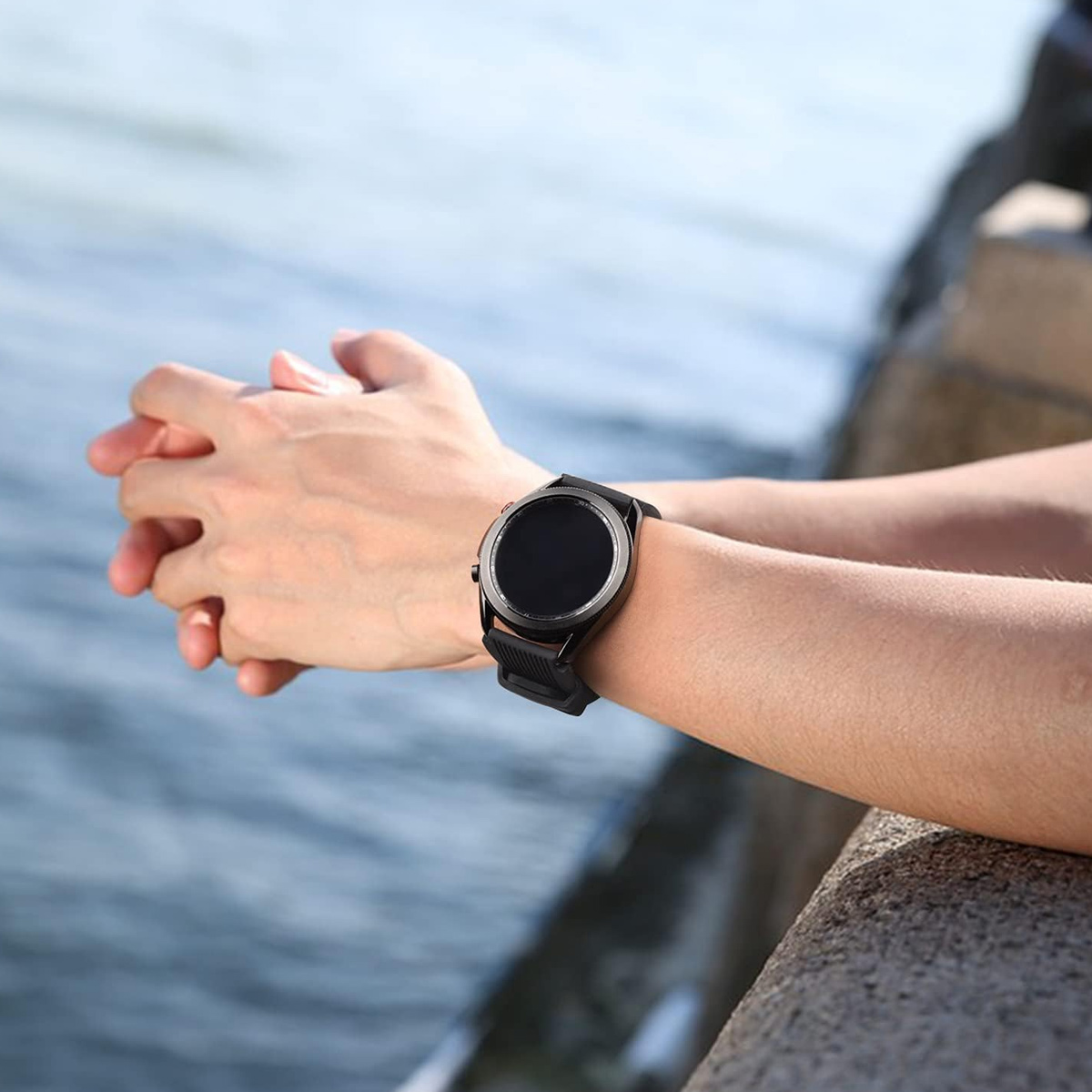 Eine Person trägt eine schwarze Smartwatch am Handgelenk, die Hände sind gefaltet. Blaues Wasser im Hintergrund.