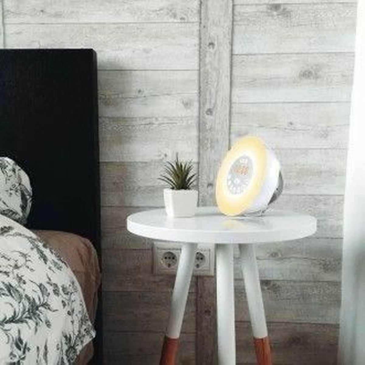 Un comodino bianco con una lampada, una piccola pianta e un letto. Il muro è di legno. La lampada è accesa.