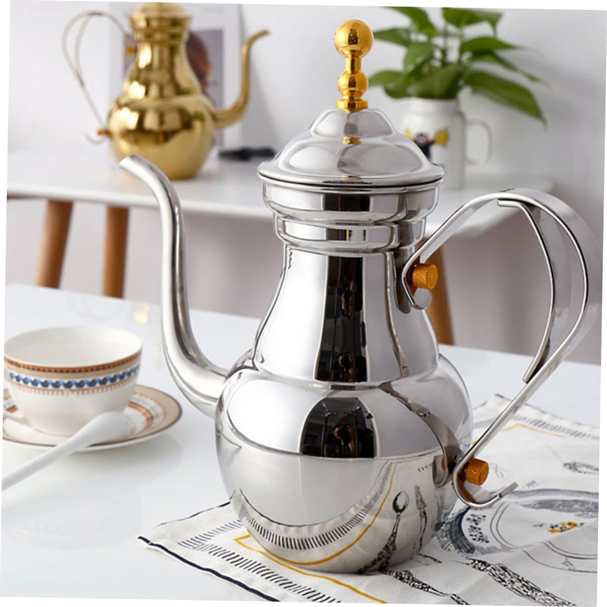 Eine glänzende silberne Teekanne mit goldenen Akzenten steht auf einem Tisch. Eine Teetasse und ein Löffel sind in der Nähe.