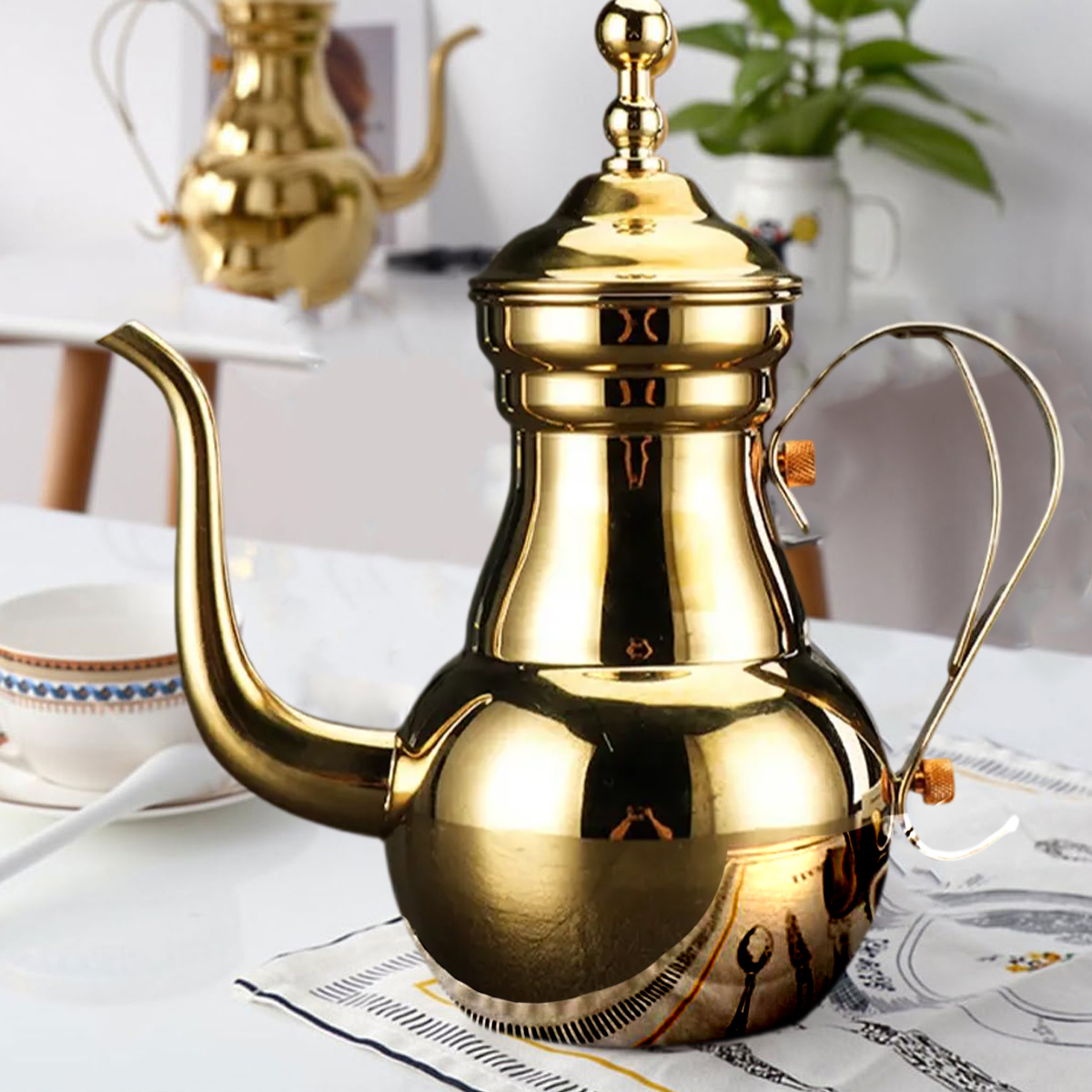 Eine goldene Teekanne steht auf einem weißen Tisch. Ein goldener Griff und Ausguss sind sichtbar. Eine weitere Teekanne befindet sich im Hintergrund.