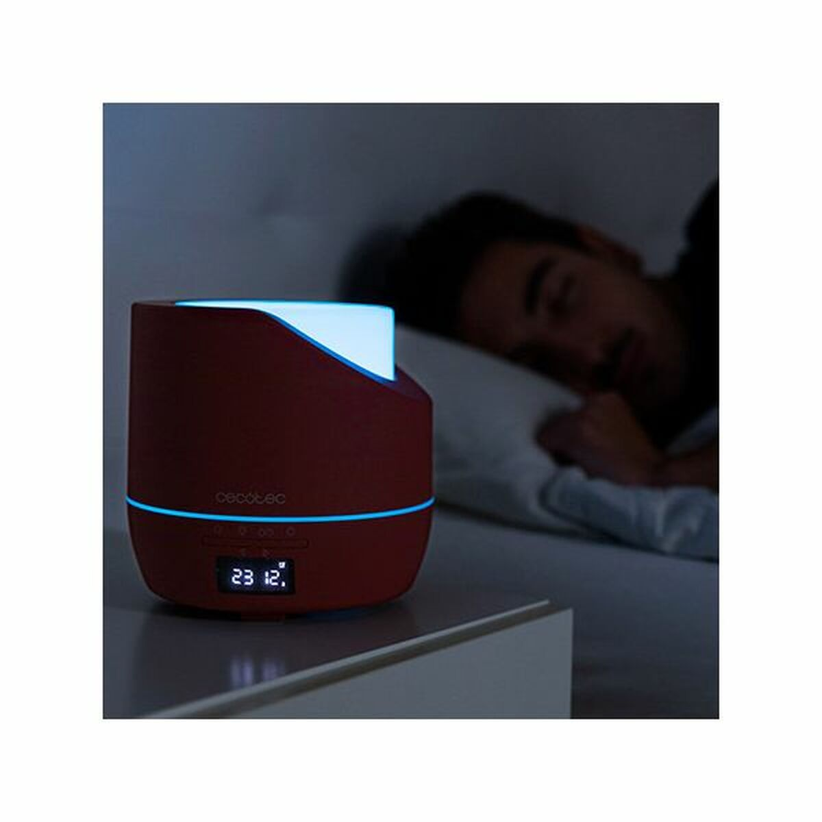 Czerwony nawilżacz z niebieskim światłem stoi na stoliku nocnym. W tle śpi osoba w łóżku.