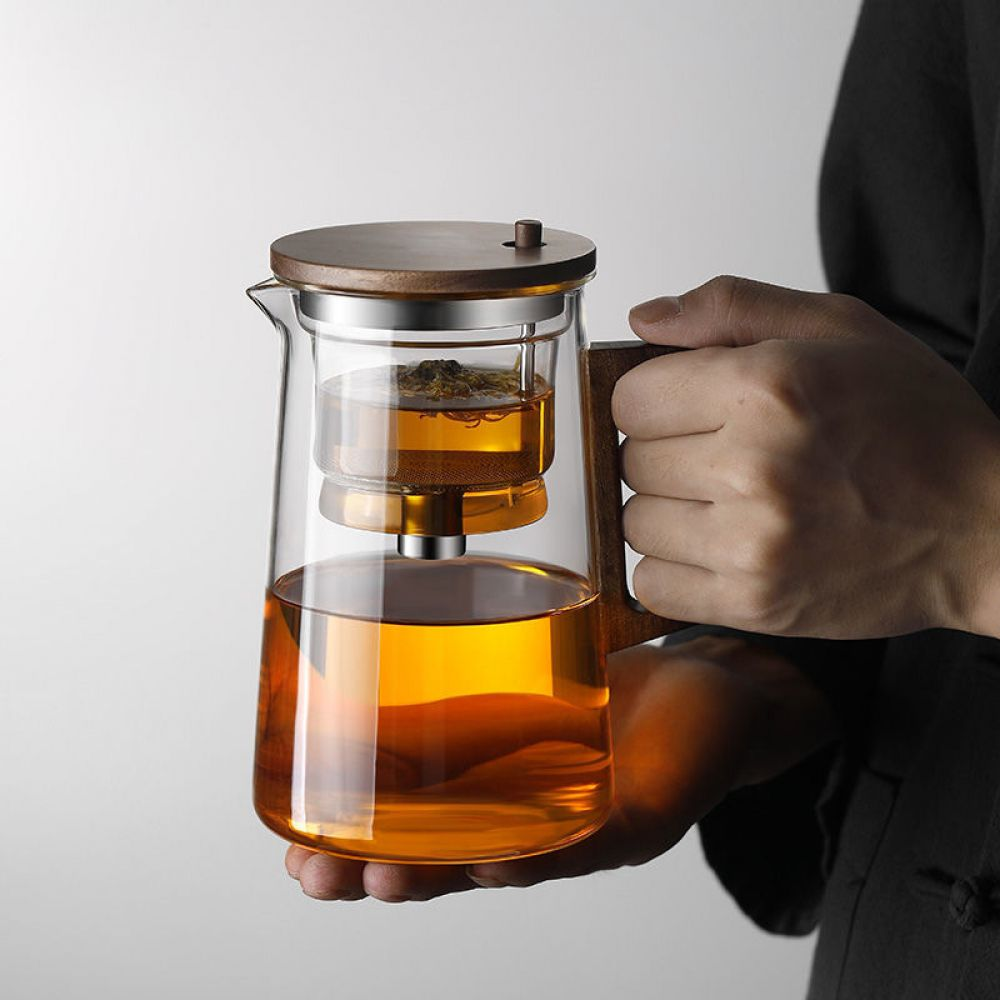 Eine Person hält eine Glasteekanne mit Holzgriff. Der Tee ist bernsteinfarben. Deckel und Griff sind aus Holz. Weißer Hintergrund.