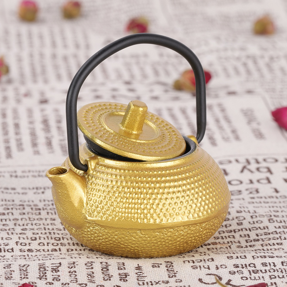 Eine goldene Teekanne mit schwarzem Griff und Deckel steht auf einer Zeitung. Kleine Blumen sind um die Teekanne verstreut.