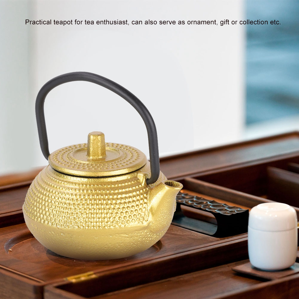 Eine goldene Teekanne mit schwarzem Griff steht auf einem Holztablett. Eine weiße Tasse, Untersetzer und ein schwarzes Utensil sind ebenfalls sichtbar.