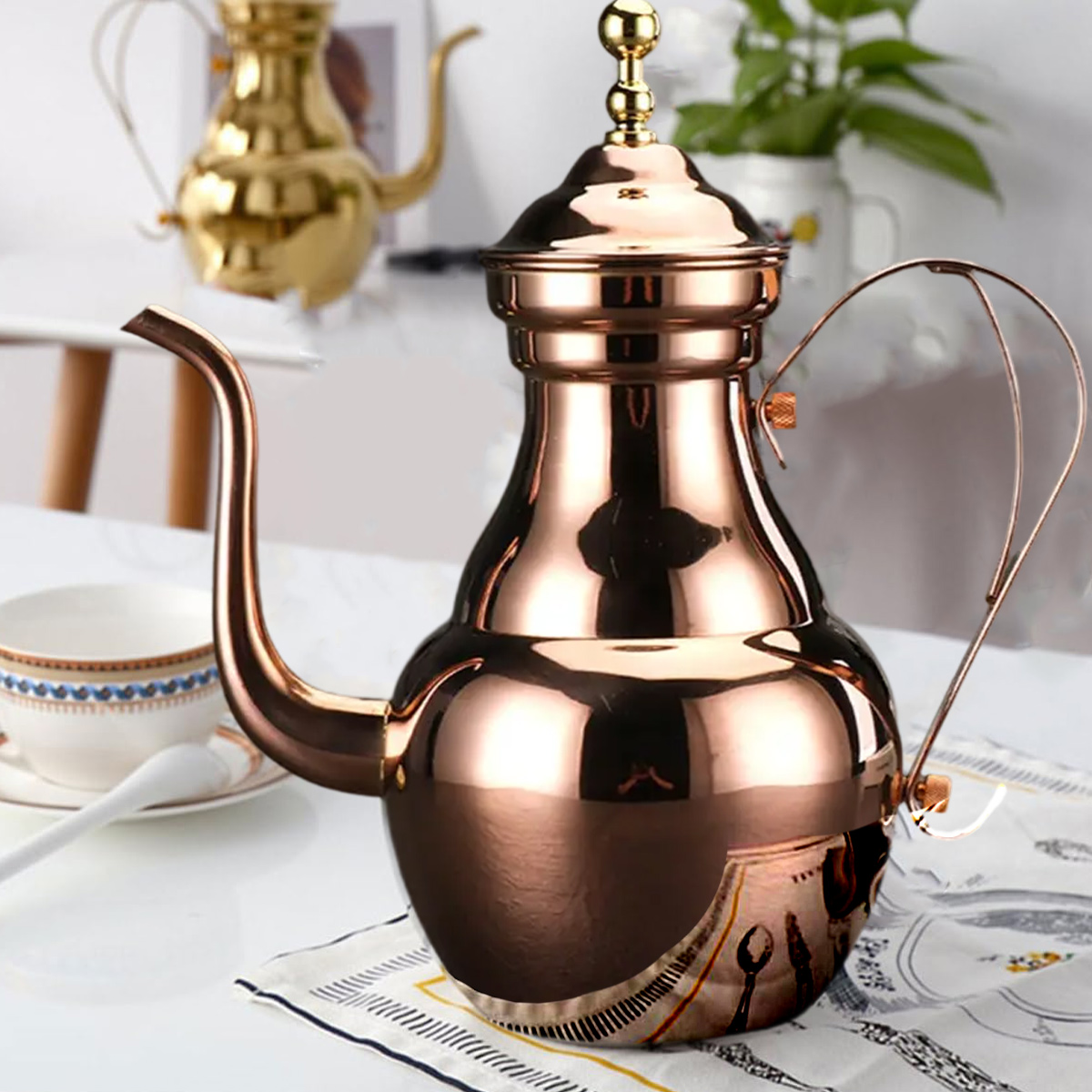 Eine Kupferkanne steht auf einem Tisch mit einer weißen Tasse und einem Löffel. Im Hintergrund steht eine goldene Teekanne.