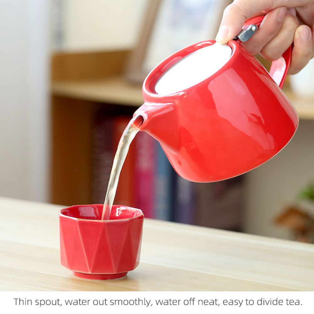 Ein roter Teekessel gießt Wasser in eine rote Tasse. Die Hand hält die Teekanne. Im Hintergrund ein Holzregal.